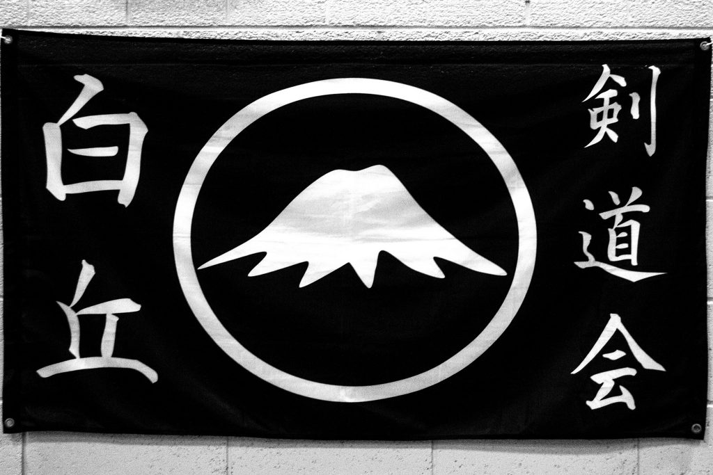 The Club Flag. The Mon is in the middle visually representing a White Hill and evoking the imagery of Mt Fuji. The Kanji to the left reads Shira (white) and Oka (hill) while the kanji on the right reads Ken (Sword) Doh (Way/Path) Kai (Association).